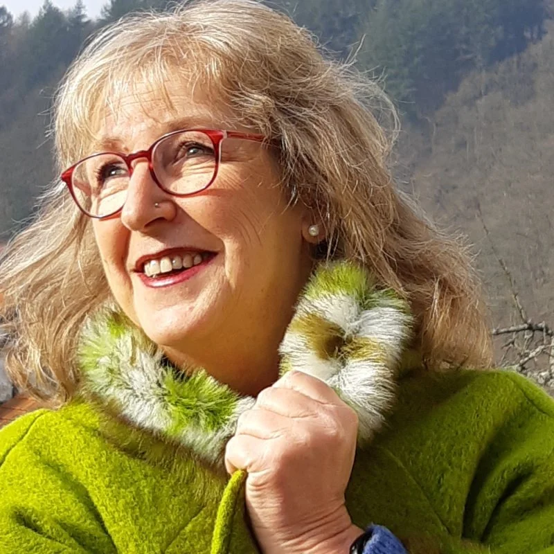 Lächelnde ältere Frau mit roten Brillen und grünem Mantel vor Bergkulisse.