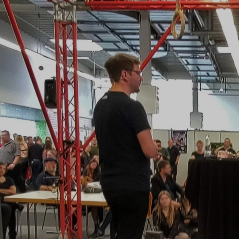 Mann spricht vor Publikum in einer Messehalle bei einer Podcast-Veranstaltung.