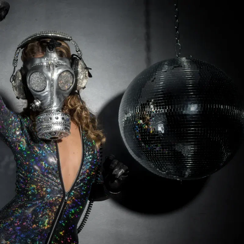 Person mit Gasmaske und glitzerndem Outfit neben einer Discokugel vor dunklem Hintergrund