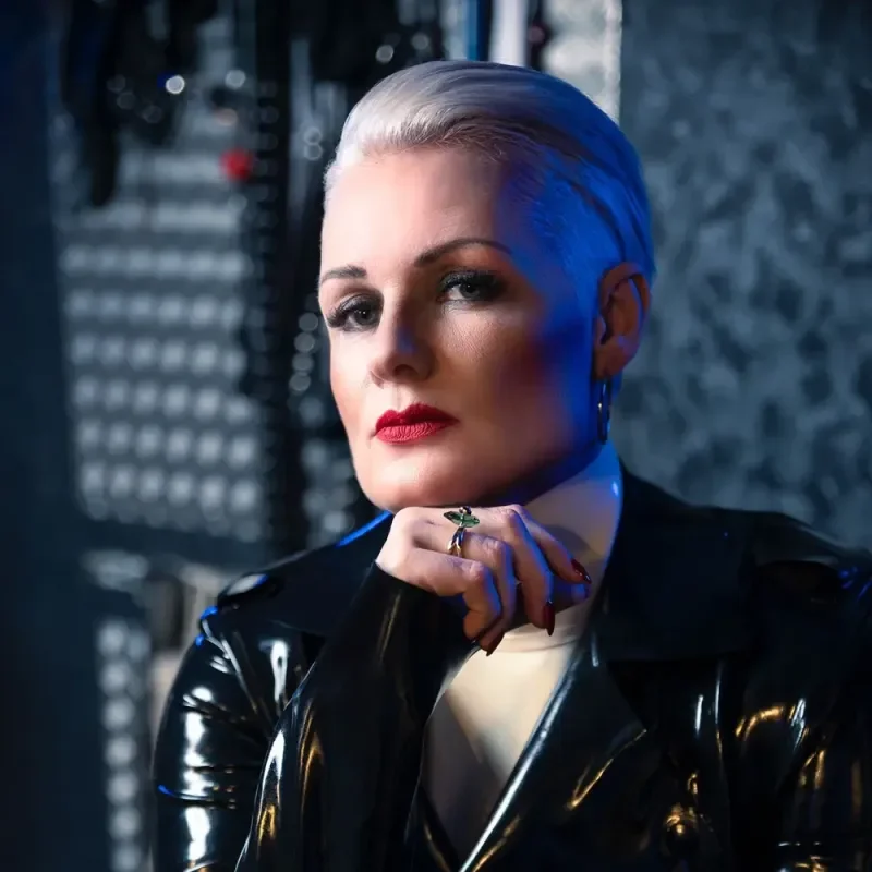 Porträt einer Frau mit platinblondem Haar, rotem Lippenstift und schwarzer Lackjacke in kühlem Licht