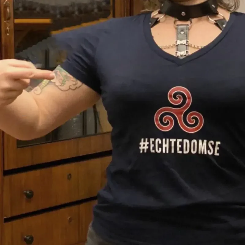 Person mit schwarzem #ECHTEDOMSE-Shirt, Halsband und Tattoo am Oberarm, auf das sie zeigt