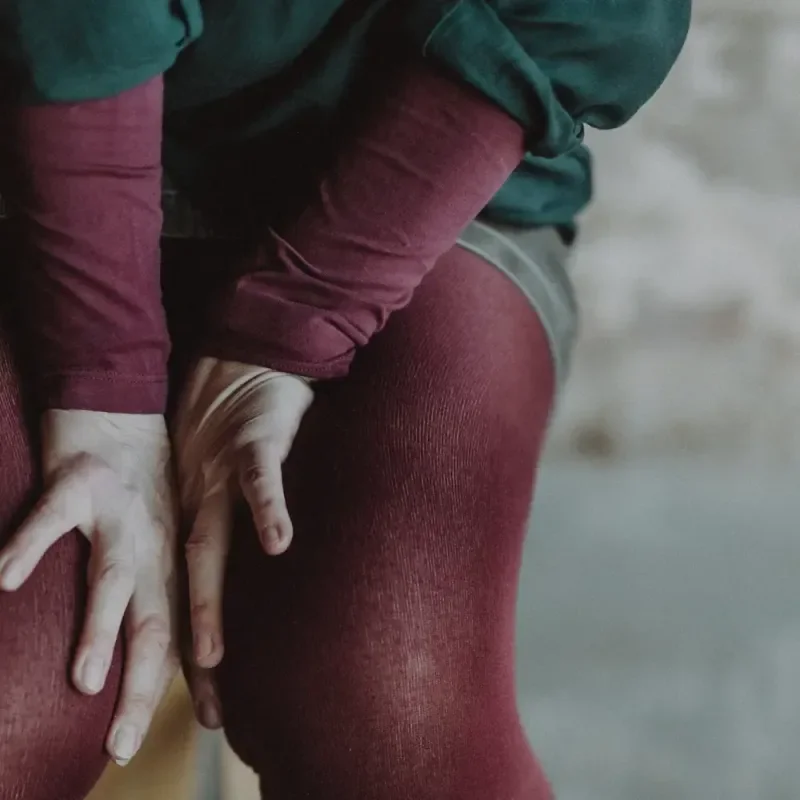Nahaufnahme einer sitzenden Person mit bordeauxroter Strumpfhose und grüner Kleidung vor unscharfem Hintergrund