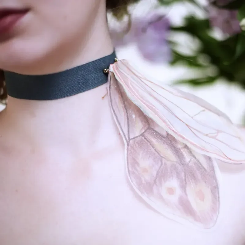 Nahaufnahme eines Halses mit schwarzem Choker und zarten rosa Feenflügeln vor blumigem Hintergrund