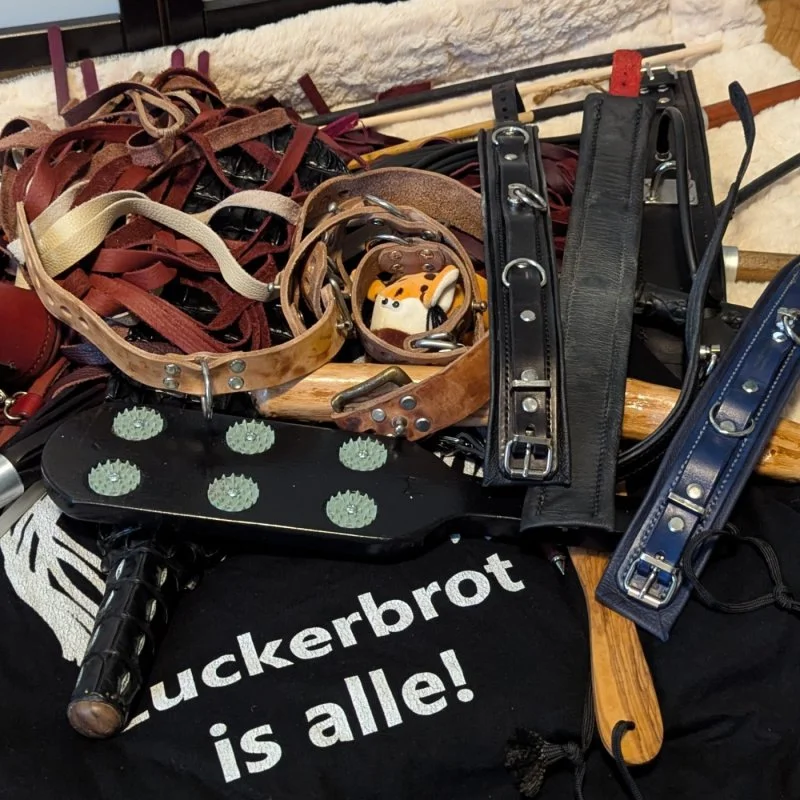 Leder- und Metall-BDSM-Accessoires auf schwarzem Stoff mit dem Schriftzug Zuckerbrot is alle!