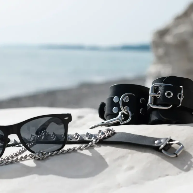 Sonnenbrille, Lederfesseln und Halsband mit Kette auf einer Mauer vor unscharfem Meereshintergrund