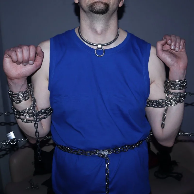 Mann in blauem Shirt mit Ketten an Armen und Taille vor dunklem Hintergrund