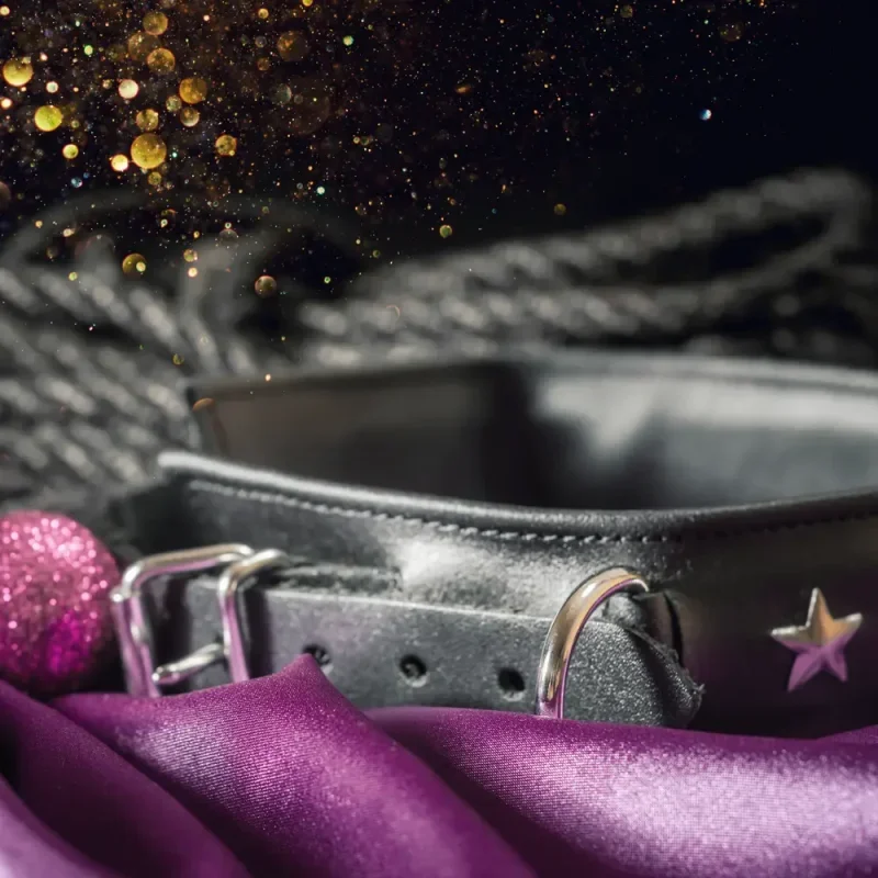 Schwarzes Halsband mit Metallringen auf violettem Stoff, umgeben von Glitzer und weihnachtlichen Dekoakzenten