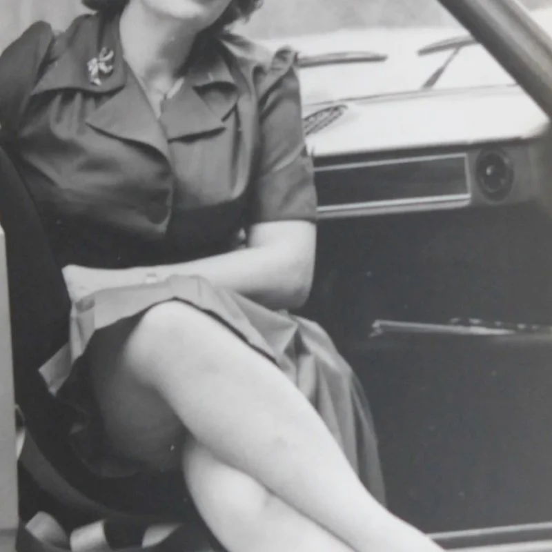 Schwarzweiß-Porträt einer sitzenden Frau in Uniform mit übereinandergeschlagenen Beinen vor einem Auto.