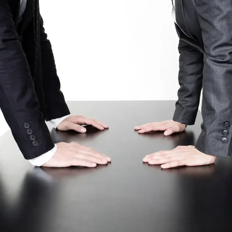 Zwei Personen in Anzügen stehen sich mit Händen auf einem schwarzen Tisch gegenüber