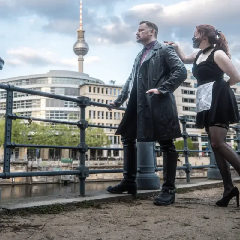 Dominanter Mann und Frau in Dienstmädchen-Outfit posieren an der Spree vor dem Berliner Fernsehturm