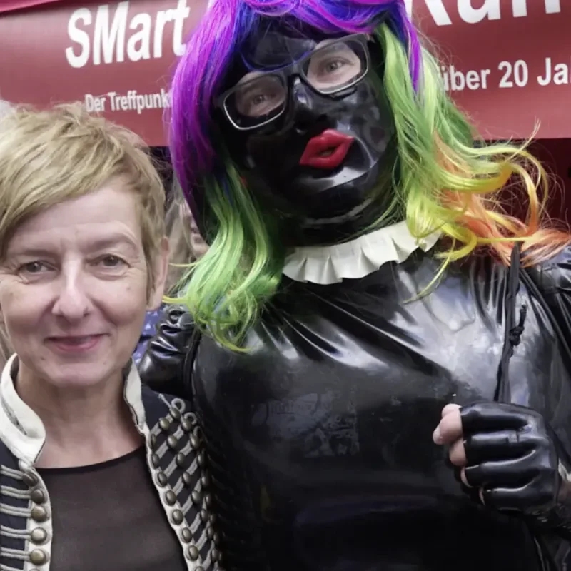 Zwei Personen vor SMart-Rhein-Ruhr-Banner, eine in schwarzem Lackoutfit mit bunter Perücke und Maske