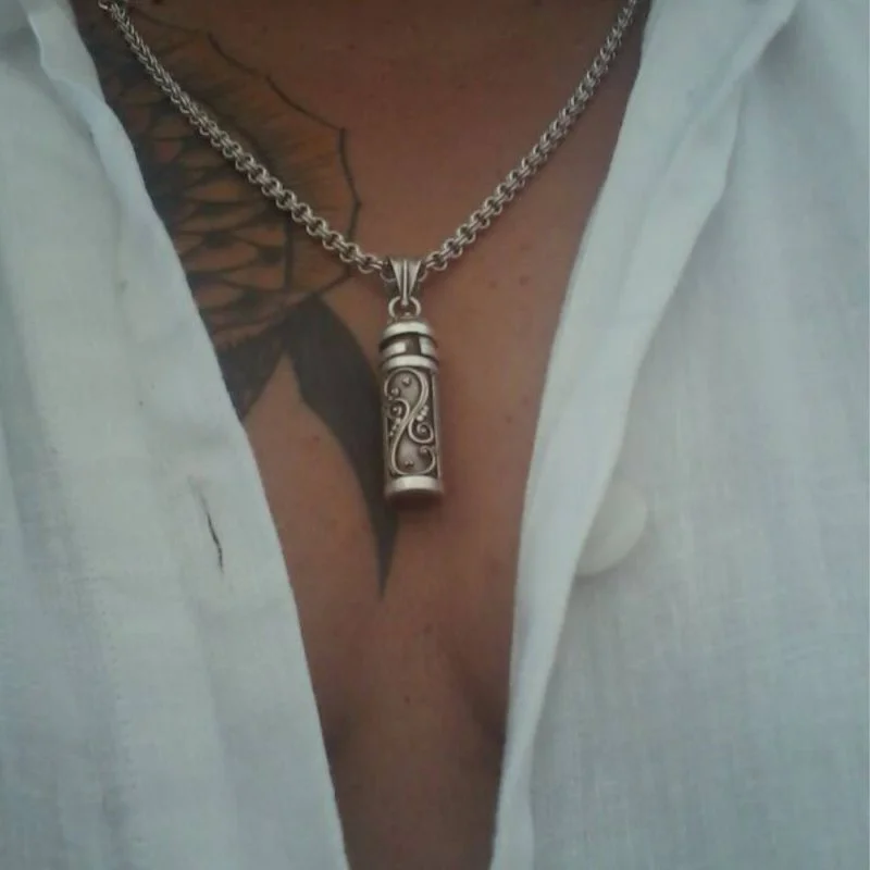 Nahaufnahme eines Oberkörpers mit offenem weißen Hemd, Kette mit Anhänger und sichtbarem Tattoo.