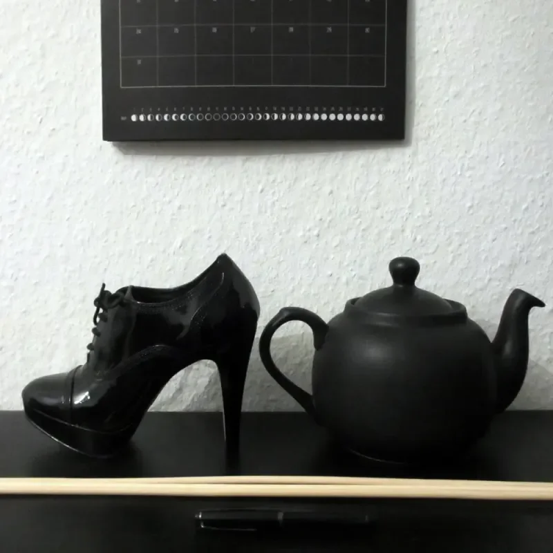 Schwarzer Lack-High-Heel neben schwarzer Teekanne auf einem Regal vor heller Wand mit Kalender im Hintergrund