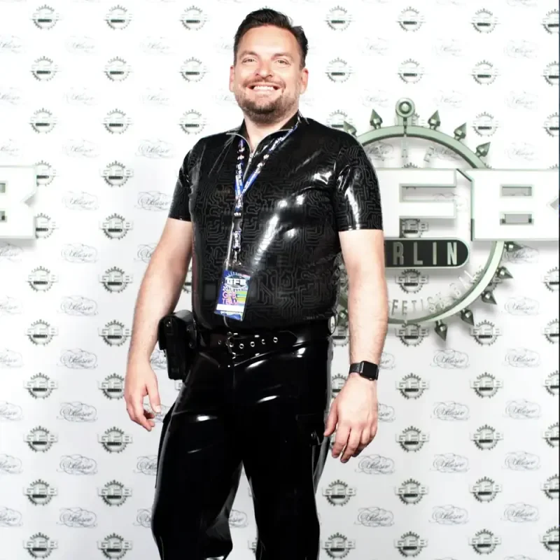Lächelnder Mann in schwarzem Lack-Outfit vor Event-Logo-Wand in Berlin