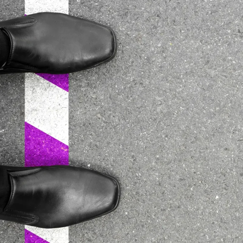 Schwarze Schuhe stehen an einer weiß-violetten Grenzlinie auf grauem Asphalt