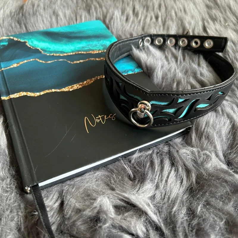 Schwarzes Halsband mit türkisfarbenem Muster auf einem Notizbuch auf grauem Fell.