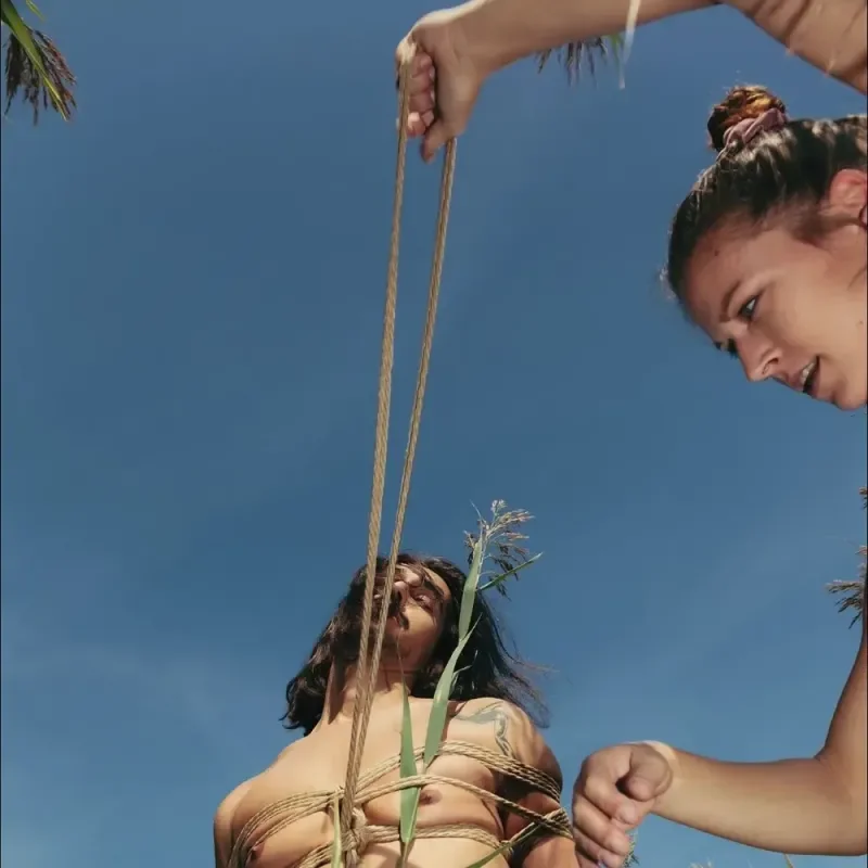 Frau bindet eine andere Frau mit Seilen unter blauem Himmel, Podcast-Cover zur Folge Shibari Sari