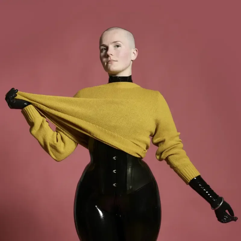 Person in gelbem Pullover, schwarzem Latexkorsett und Handschuhen vor rosa Hintergrund