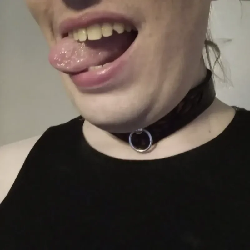 Nahaufnahme eines Mundes mit herausgestreckter Zunge, schwarzem Halsband mit Ring und schwarzem Oberteil