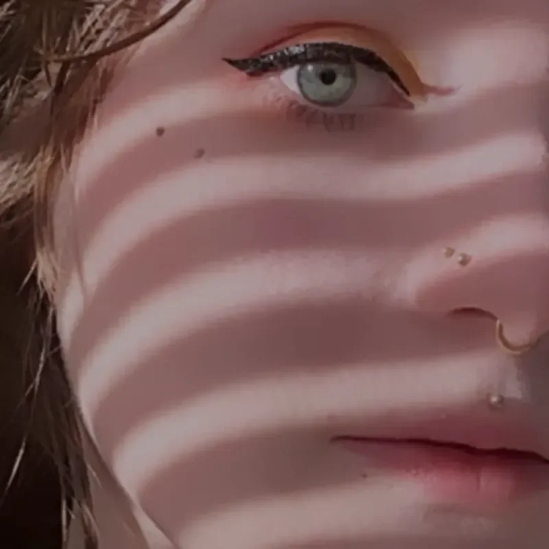 Nahaufnahme eines Gesichts mit Piercings, markantem Eyeliner und gestreiftem Schattenmuster über der Haut