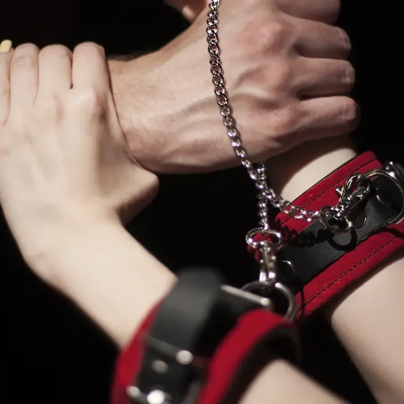 Nahaufnahme gefesselter Handgelenke mit roten Manschetten und Kette im BDSM-Kontext