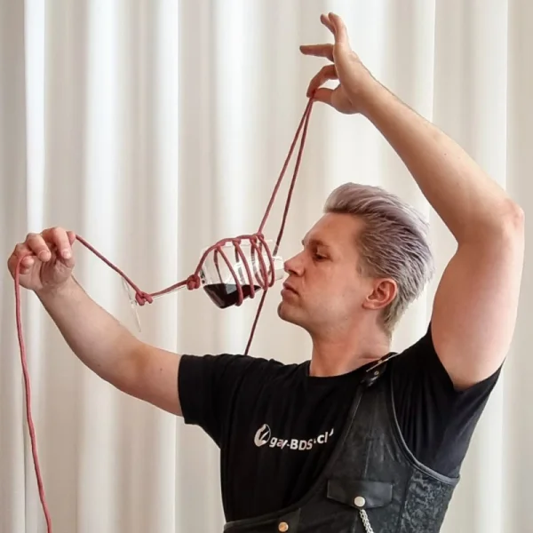 Person mit erhobenen Armen hält eine rote Schnur mit einem kleinen Objekt vor hellem Vorhang.