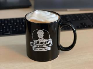 Schwarze Tasse mit Podcast-Logo und aufgeschäumtem Getränk vor Laptop und Tastatur auf dem Schreibtisch