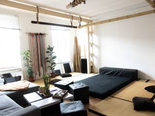 Heller Wohn- und Studioraum mit Tatami-Matten, schwarzem Sofa, niedrigen Tischen, Pflanzen und großer Liegefläche.