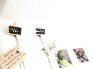 Kleine Klammern mit beschrifteten Zetteln und Fotos hängen an Schnüren vor einer hellen Wand.