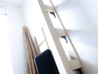 Eine Holzleiter lehnt in einem hellen Raum an einer weißen Wand, daneben stehen Holzstangen und ein schwarzer Gegenstand.
