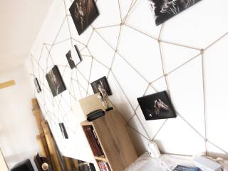 Helle Wand mit geometrischem Fadennetz, daran befestigten Fotos und einem kleinen Regal neben einem Bett.